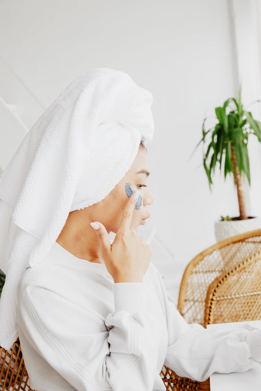 Woman applying moisturizer on her face after shower, wearing a towel on her head and white robe